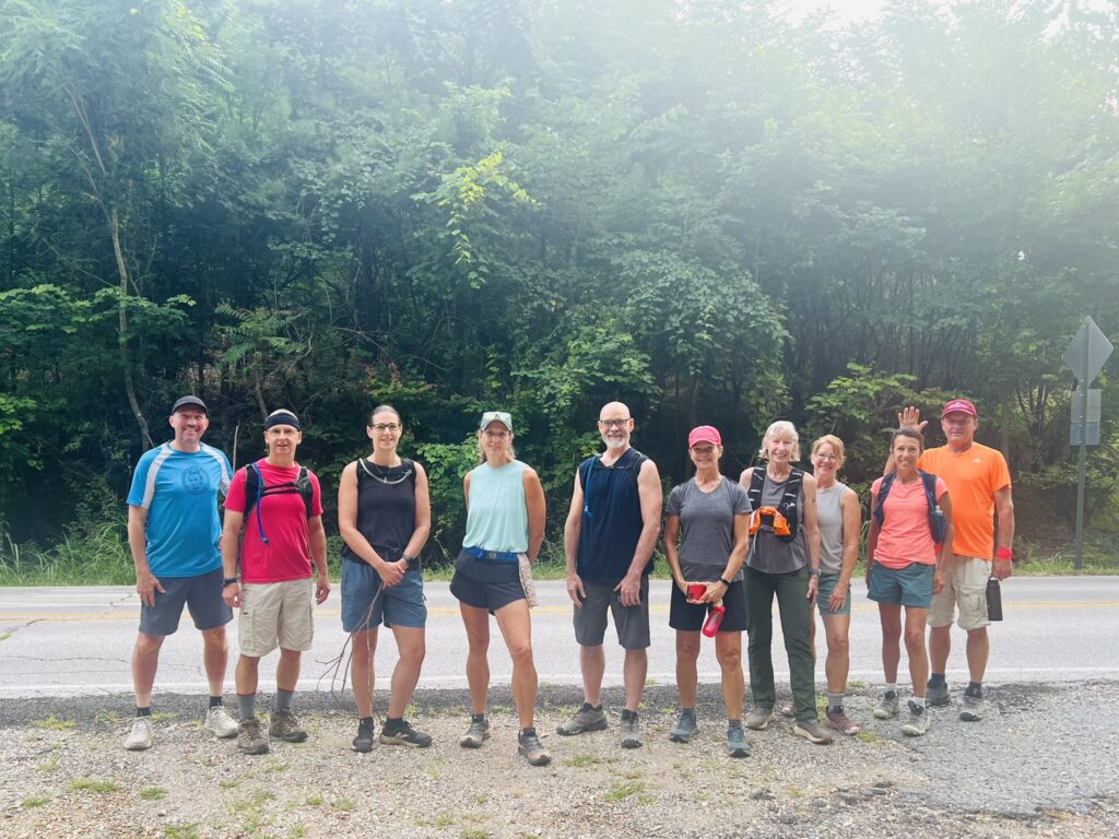 Ten hikers in T-shirts and shorts stand together along a road in front of green summer woods
