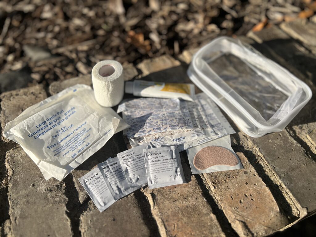 The contents of a day-hike first-aid kit laid out on a brick path, including gauze, a bandage wrap, ointment, wipes, moleskin, and a clear zipper bag