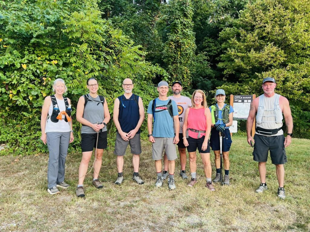 Nine hikers in summer gear stand together in the grass beside a trail map sign at the edge of the woods