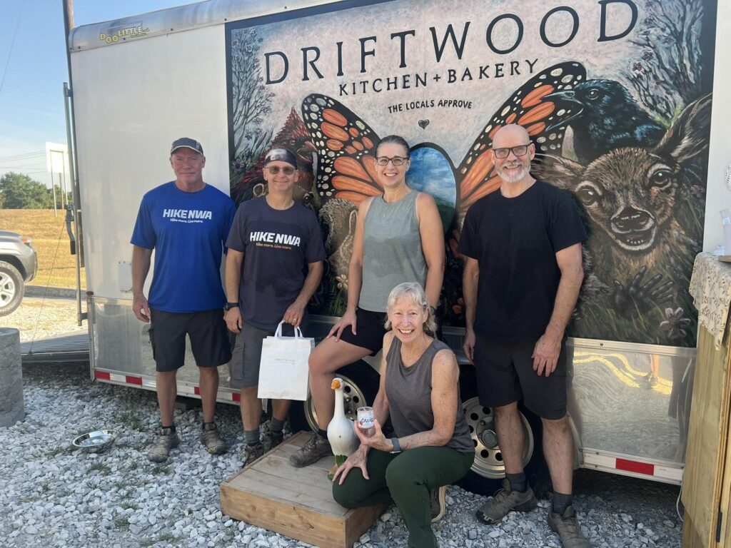 Five smiling hikers, two in Hike NWA shirts, pose in front of a food trailer painted with butterflies and animals