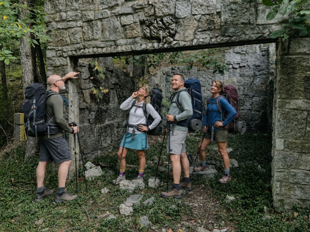 Four hikers wearing daypacks standing beside an old stone wall in the woods
