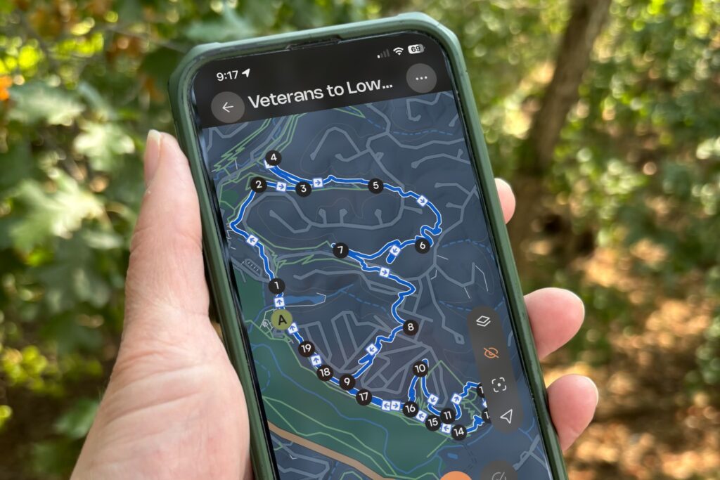 A hiker holds up a phone showing a Hike NWA route in the Komoot app, with woods behind