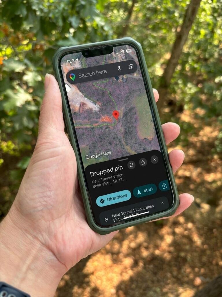 A hand holding a phone showing a satellite map with a red dropped pin and location details at the bottom of the screen