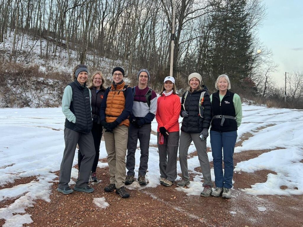 Seven hikers in winter jackets and hats stand together on a snowy gravel lot at the edge of the woods